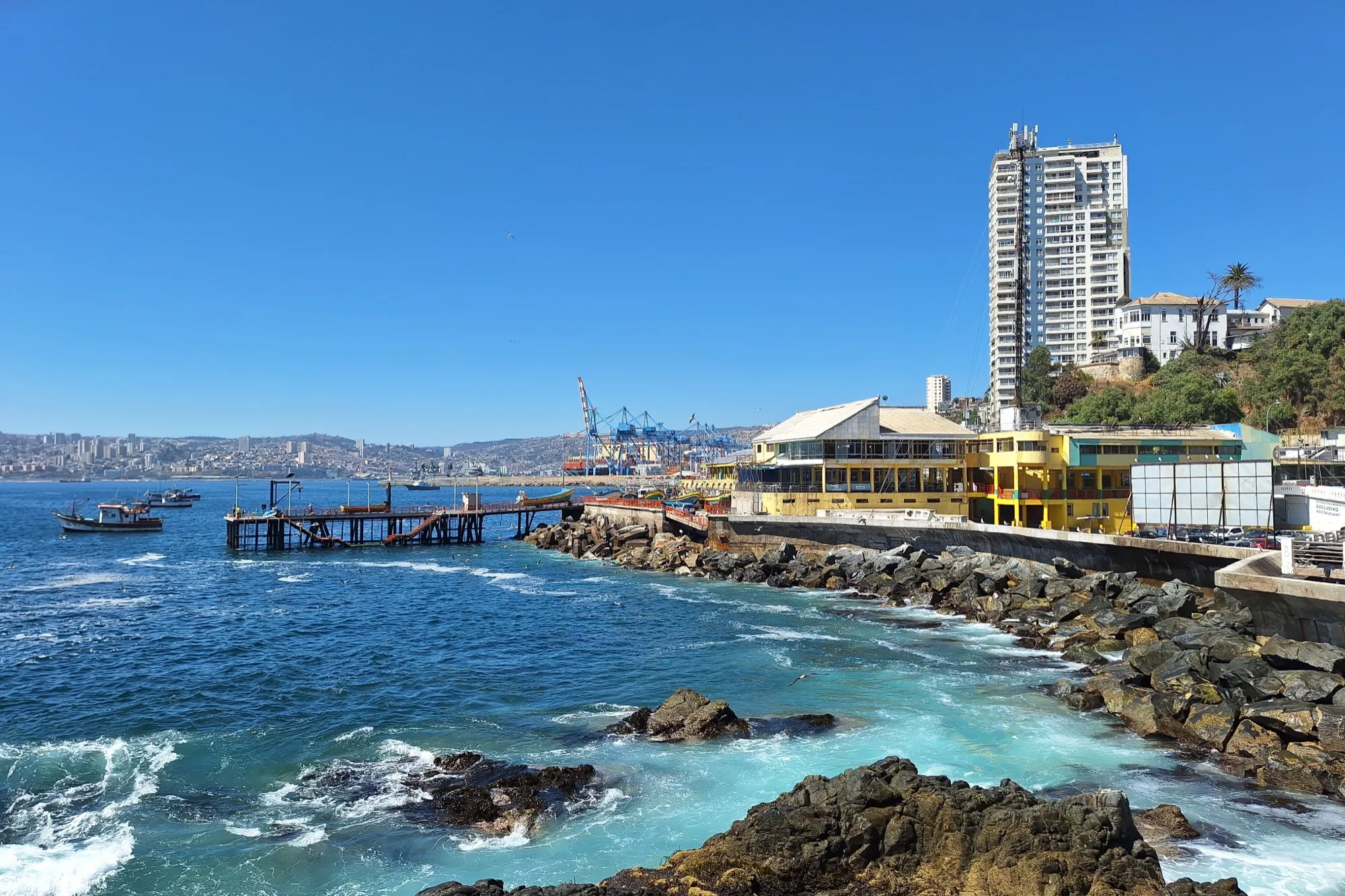 Costa rocosa de la caleta El Membrillo, con un muelle de madera, un restaurante junto al mar y un edificio alto al fondo