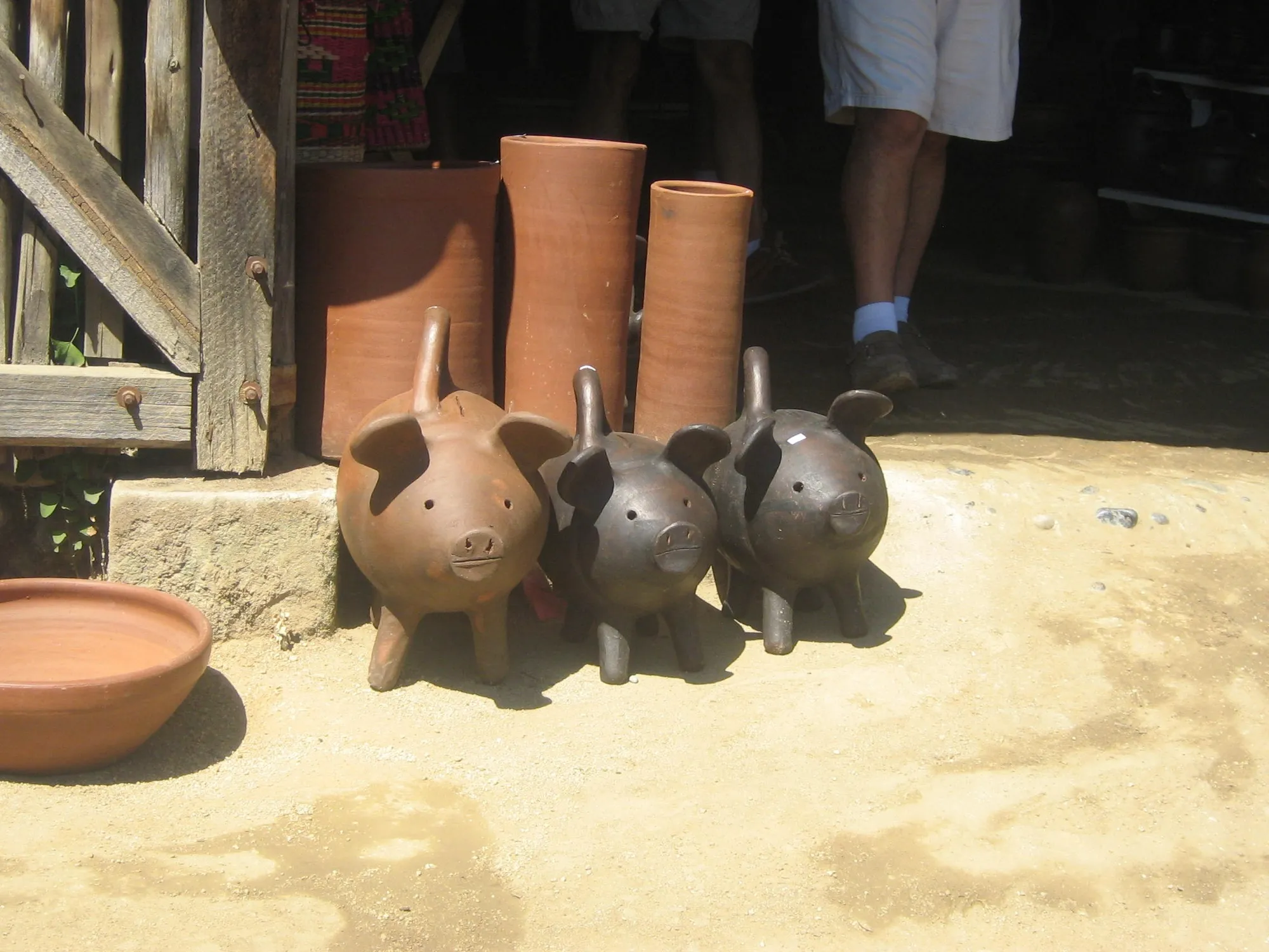 Tres chanchitos de greda de distinto color frente a un taller, con ollas de barro detrás