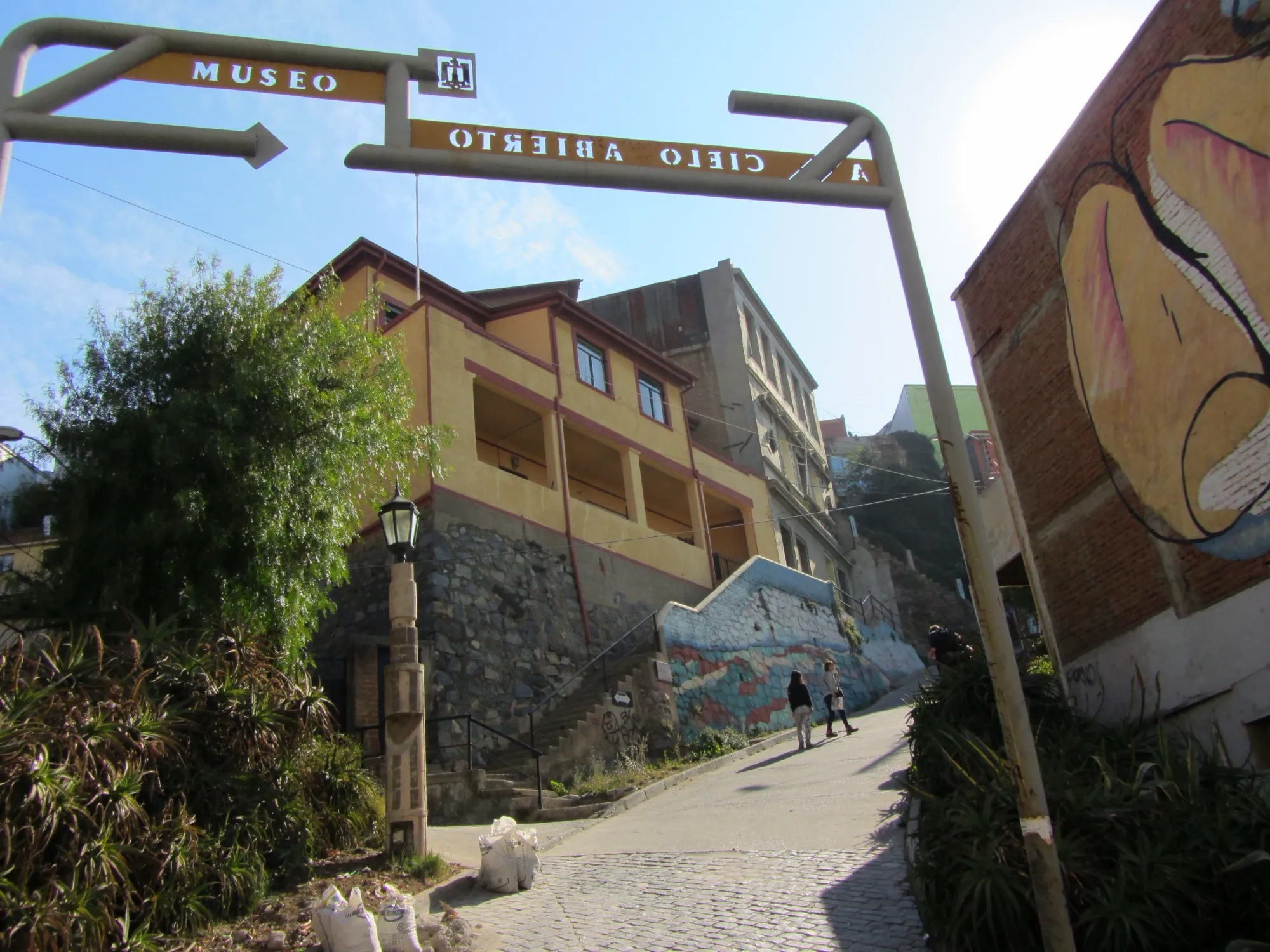 Calle empedrada que sube entre casas, con un arco metálico de señales del Museo a Cielo Abierto y un mural a la derecha