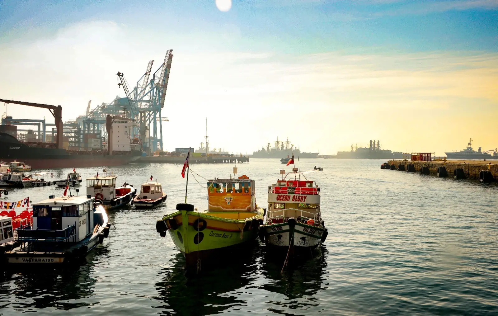 Pequeñas lanchas de colores amarradas en el puerto de Valparaíso, con grúas y barcos grises al fondo